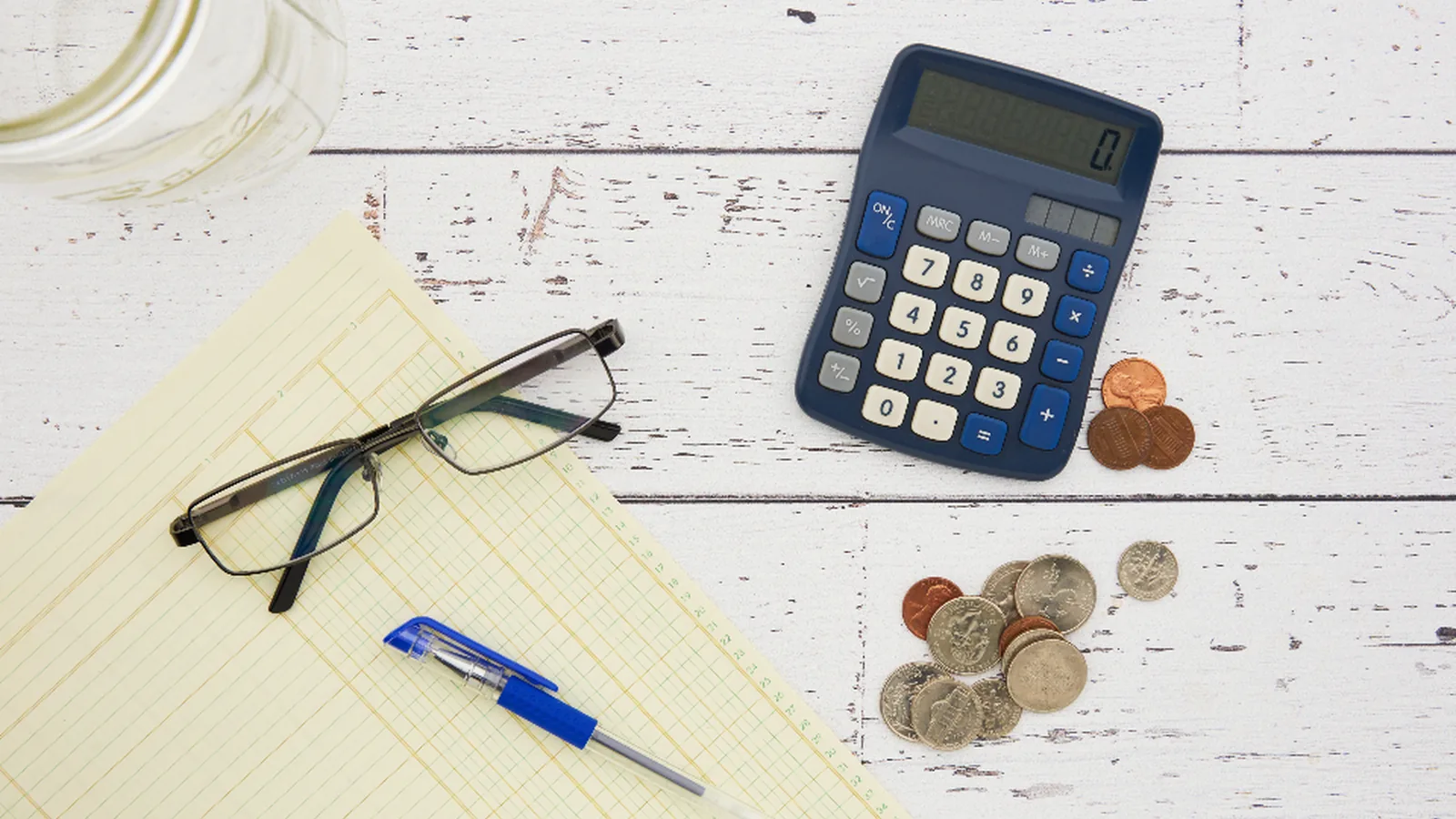 A calculator, coins, notepad and glasses on a white desk